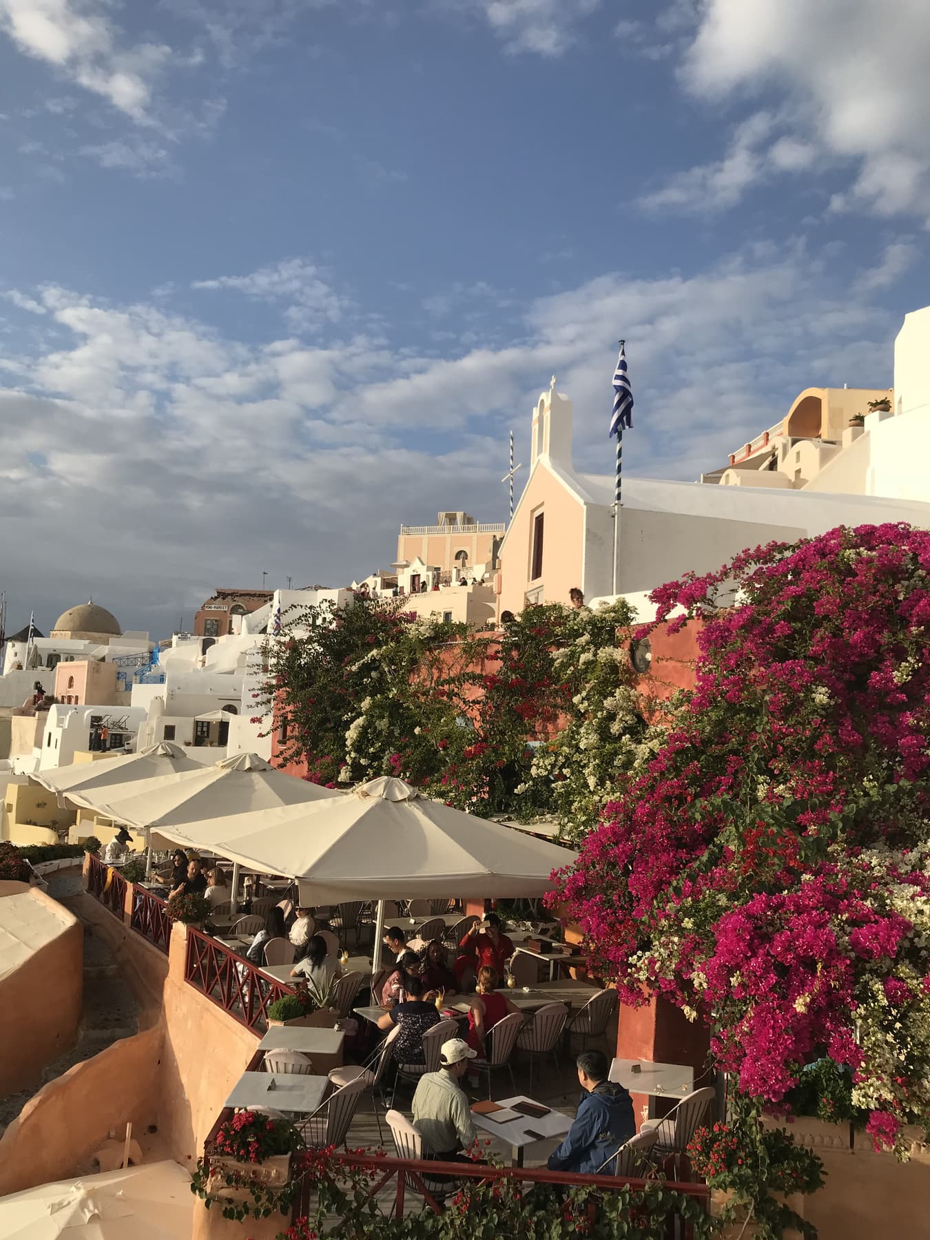 A shaded Greek terrace framed by bougainvillea, tables set out under white umbrellas